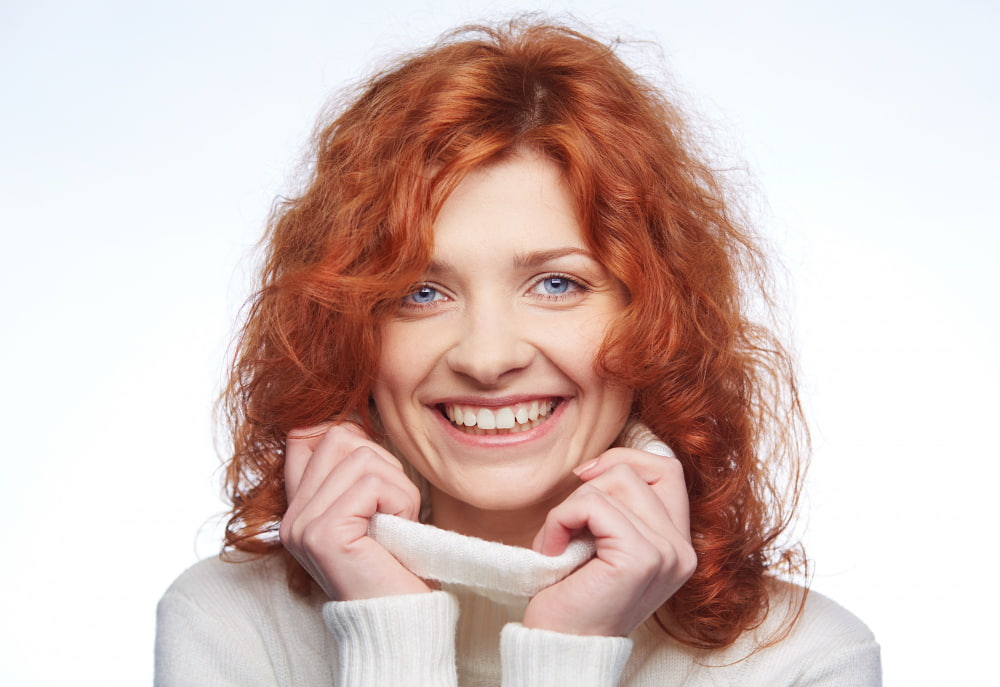 Smiling person with curly red hair, holding a white sweater collar close to their face, conveying warmth and happiness against a bright background.