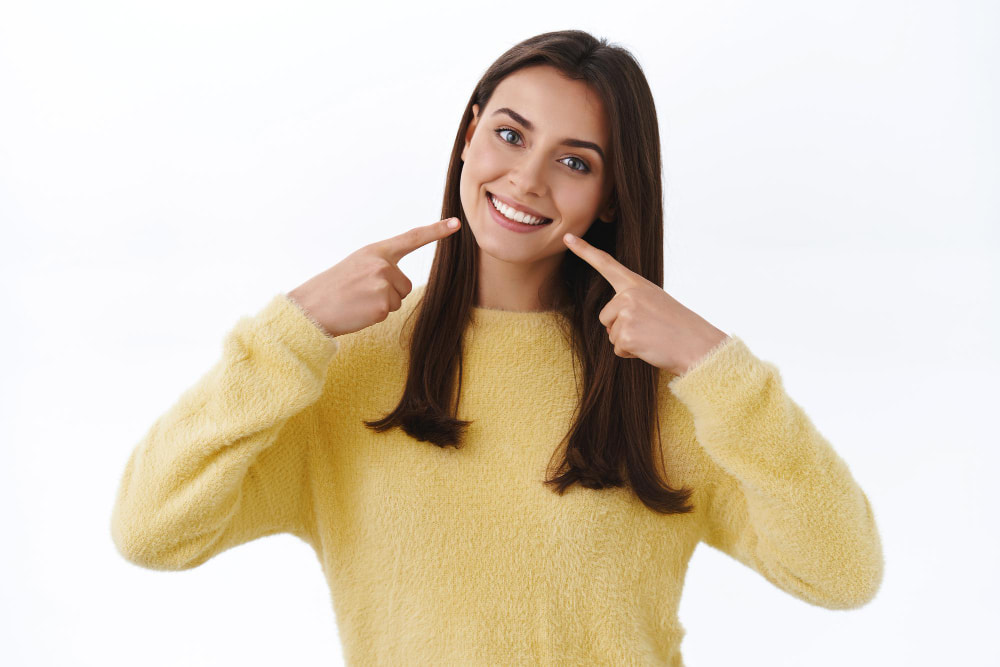 A smiling woman in a yellow sweater points at her cheeks, highlighting her bright smile.