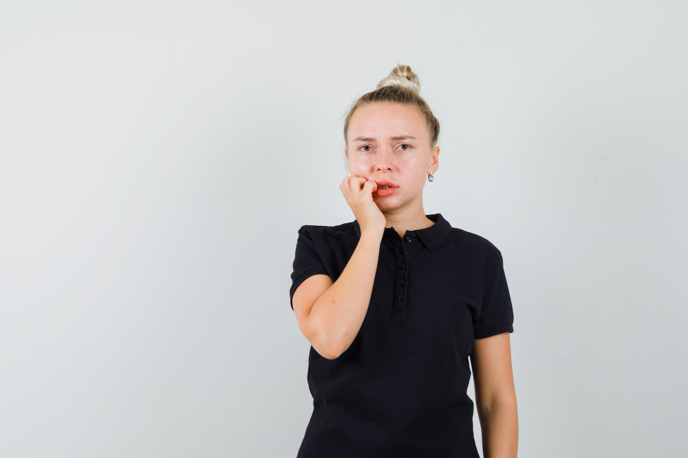 A worried person stands against a plain background, biting their fingernail and wearing a black polo shirt.