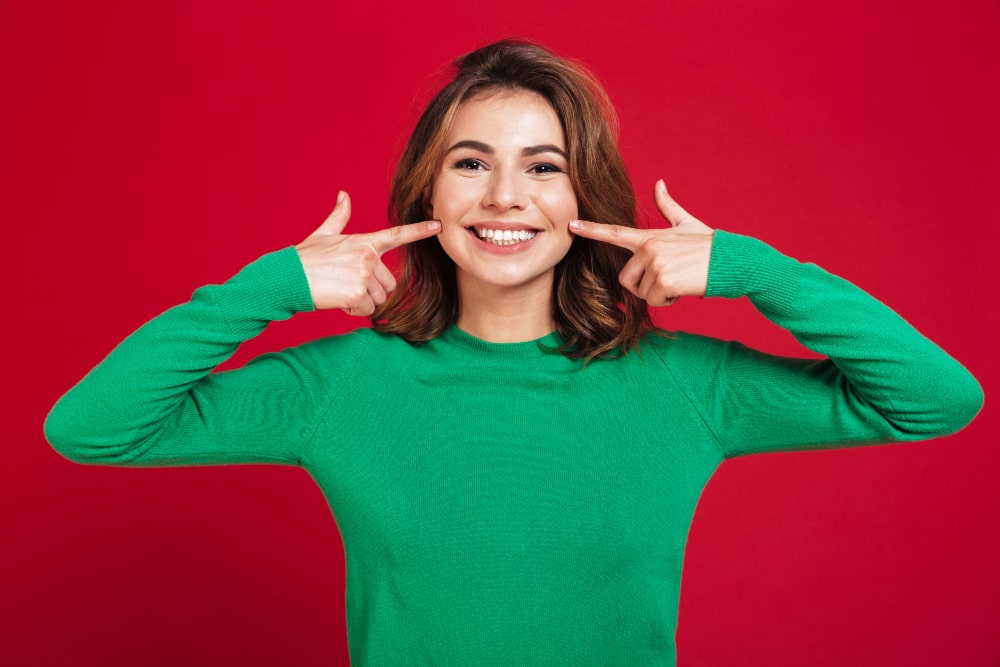 Smiling woman wearing a green sweater points to her cheeks with both index fingers against a vibrant red background.