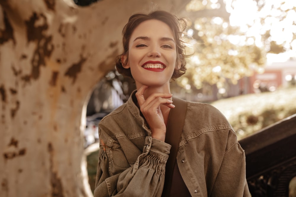 Smiling woman in a green jacket stands outdoors by a sunlit tree. Autumn leaves and a blurred park background create a warm, cheerful atmosphere.