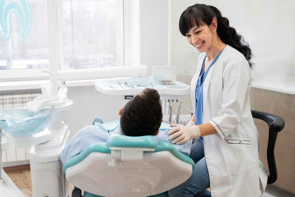 A dentist in a white coat and gloves smiles warmly at a patient reclining in a dental chair. The setting is clean, bright, and professional.