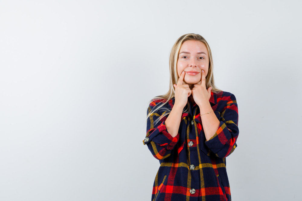 A person in a plaid shirt smiles softly, touching their cheeks with their fingers against a plain background.