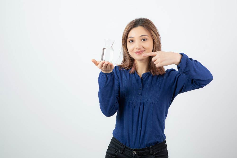 A woman in a blue shirt smiles, holding a glass of water in one hand and pointing at it with the other against a plain white background.