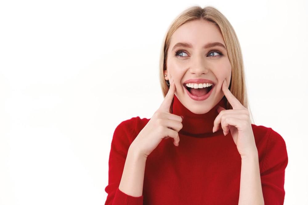 A woman in a red sweater smiles broadly, pointing at her cheeks with both index fingers.