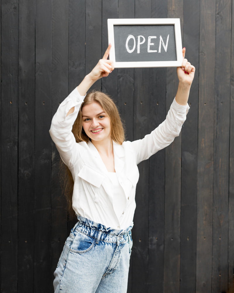 A woman in a white blouse and jeans holds a small chalkboard with "OPEN" written on it against a dark wooden background, smiling joyfully.