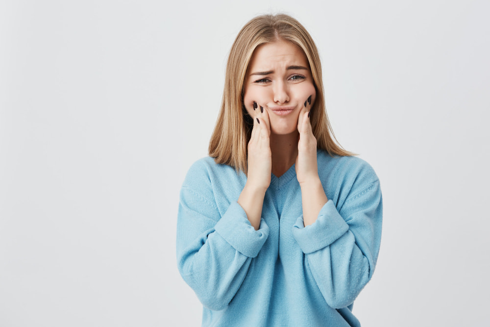 A woman in a blue sweater makes a humorous, exaggerated pout, pressing her cheeks with her hands.