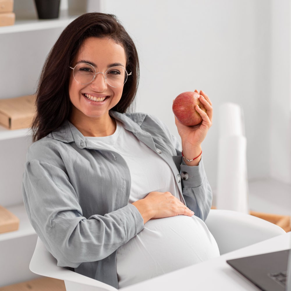 Smiling pregnant woman in glasses holds an apple while sitting in an office chair. She rests a hand on her belly.