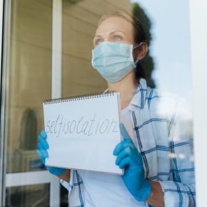 Woman wearing a face mask and gloves holding a 'self-isolation' sign while standing behind a glass door.