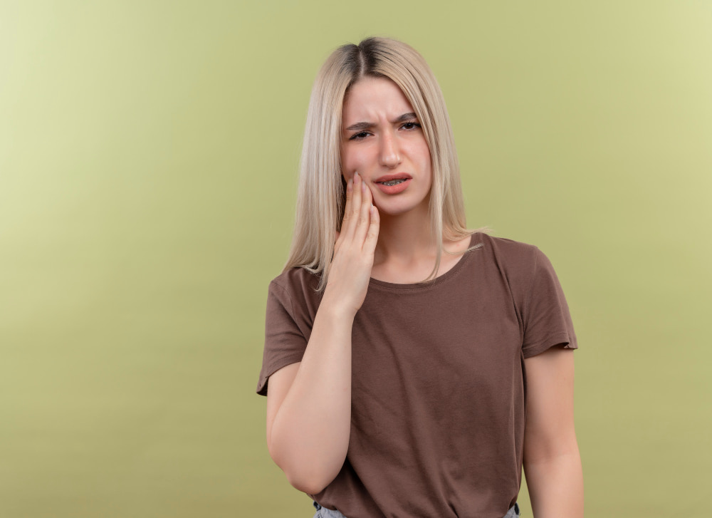 A woman with long blonde hair, wearing a brown shirt, touches her cheek with a pained expression, suggesting toothache against a soft green background.