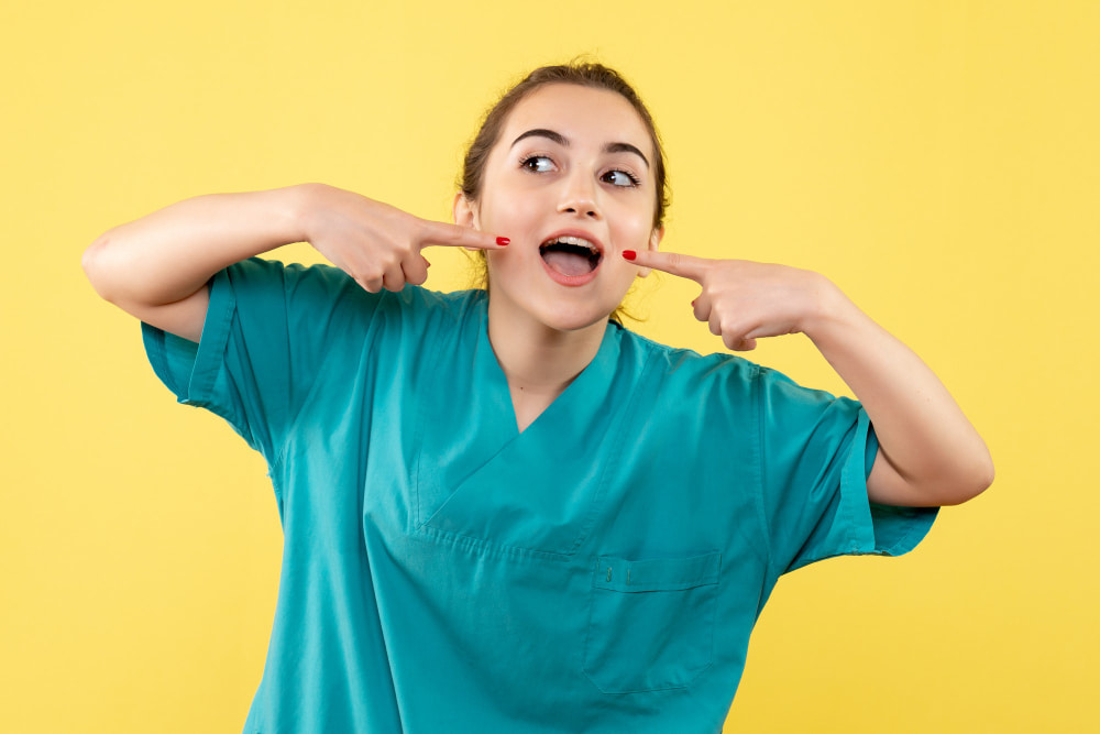 A woman in teal scrubs playfully points to her cheeks with both index fingers. She has an open mouth, expressing cheerfulness against a bright yellow background.