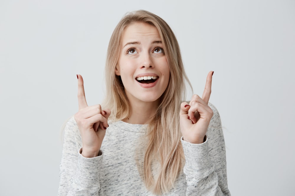 Young woman with long blonde hair looks upward with excitement, smiling and pointing up with both hands.