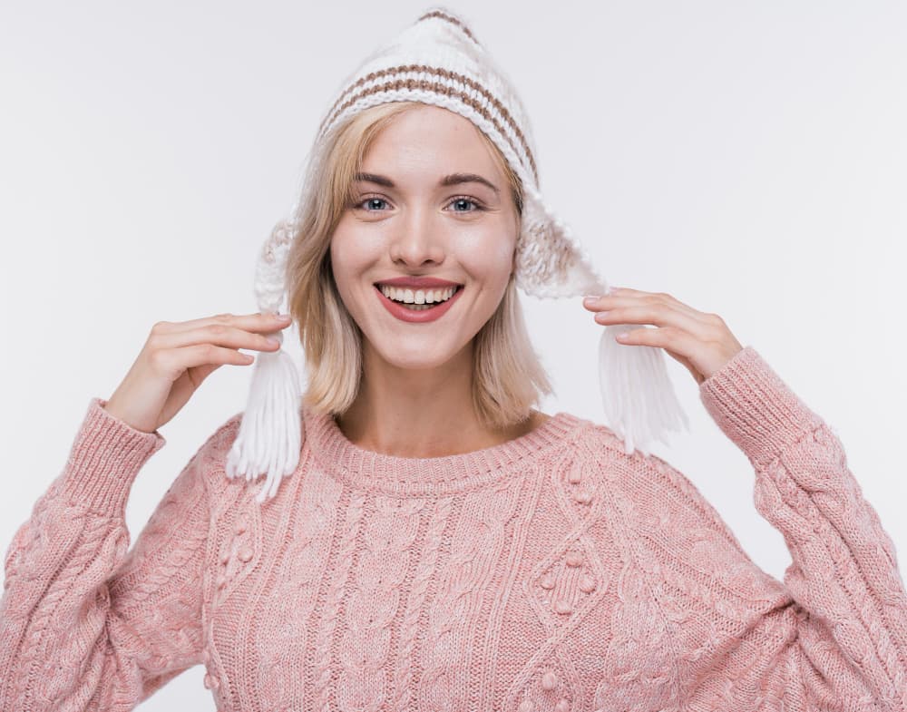 Smiling woman in a pink knit sweater and white winter hat with tassels, against a light background. She exudes warmth and happiness.