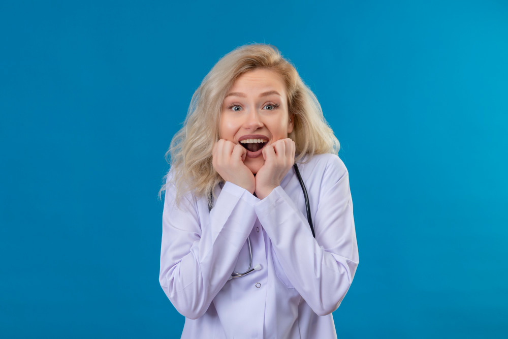 A woman with blonde hair in a white lab coat displays excitement with wide eyes and hands near her face.