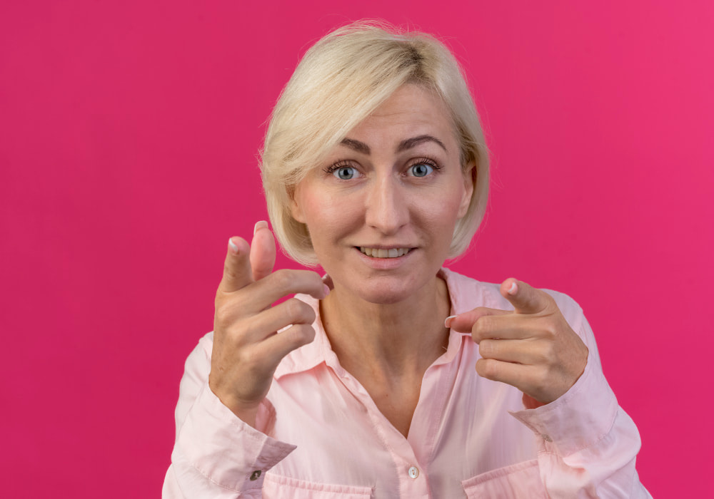 A woman with short blonde hair in a light pink shirt smiles warmly and gestures with both hands, pointing at the camera.