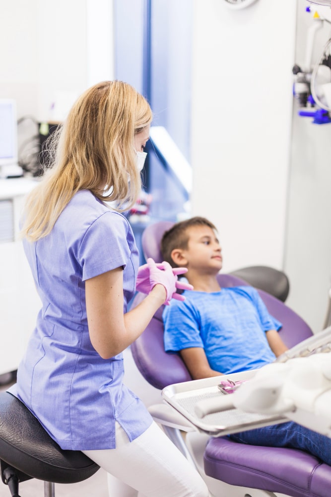 A dentist wearing gloves and a mask talks to a young boy in a blue shirt sitting in a dental chair.