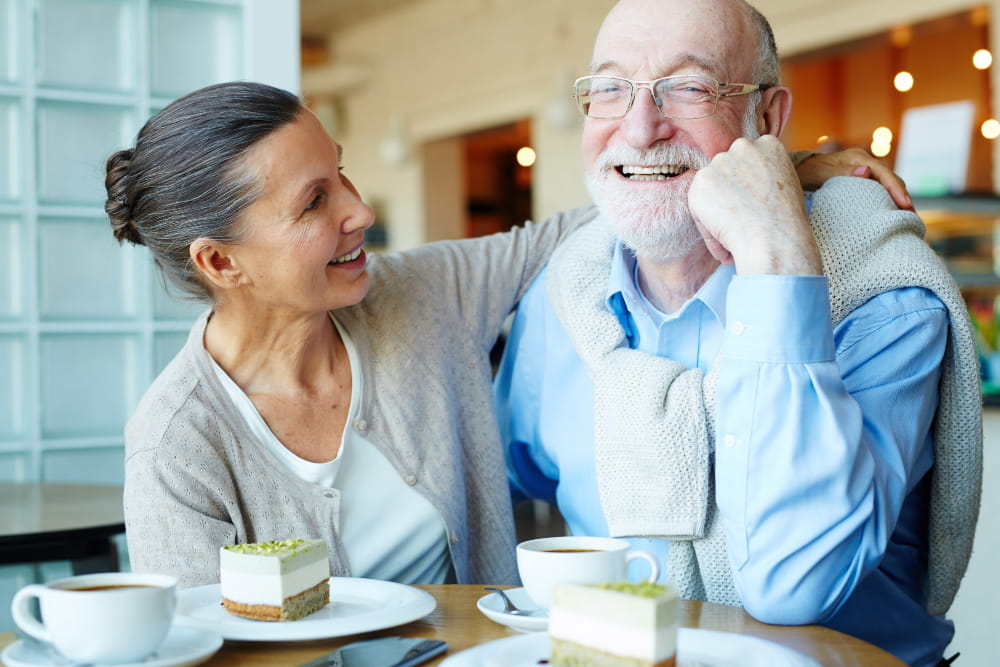 Elderly couple smiling at a cafe. The woman has her arm around the man, both look happy.