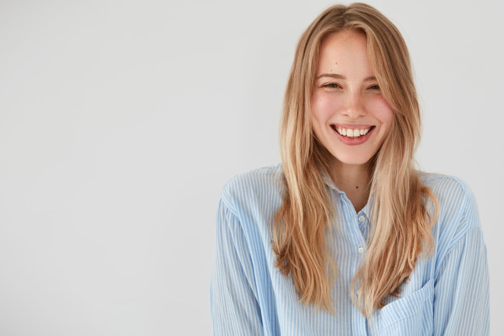 A young woman with long blonde hair smiles warmly, wearing a light blue striped shirt.