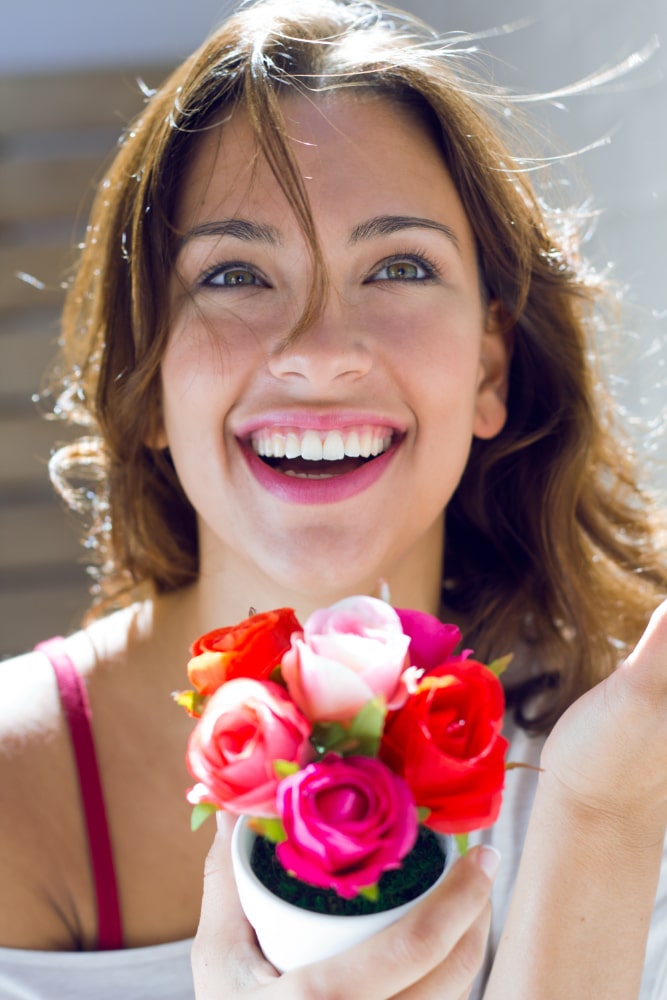 Smiling woman with brown hair holds a small bouquet of vibrant roses in reds and pinks. Bright lighting highlights her joyful expression.