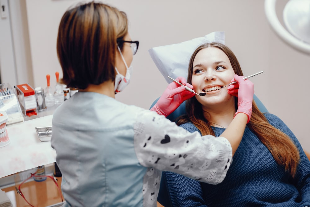 Dentist in pink gloves examines a smiling woman in a dental chair, using instruments. The scene is bright and clinical.