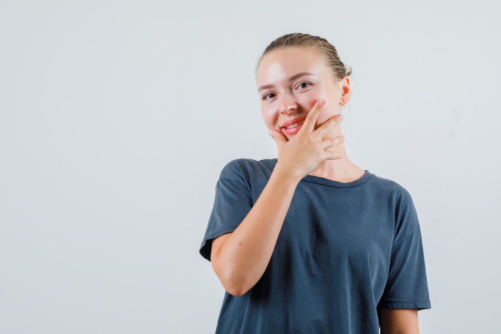 A young person in a casual blue shirt stands against a plain white background, smiling with their hand playfully covering part of their mouth,