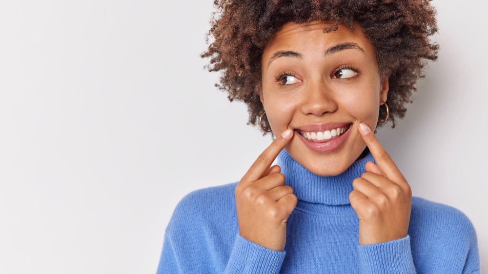 A woman in a blue sweater smiles widely, pointing at her dimples. Her curly hair and playful expression convey a joyful and cheerful mood.