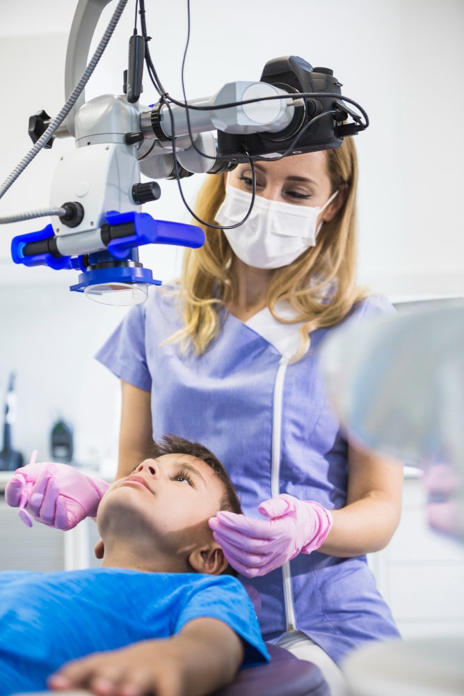 A dentist in a mask checks a young boy's teeth using a dental microscope. The dentist wears pink gloves.