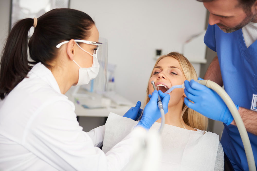 A dentist and assistant, both in protective gear, examine a patient in a dental chair.