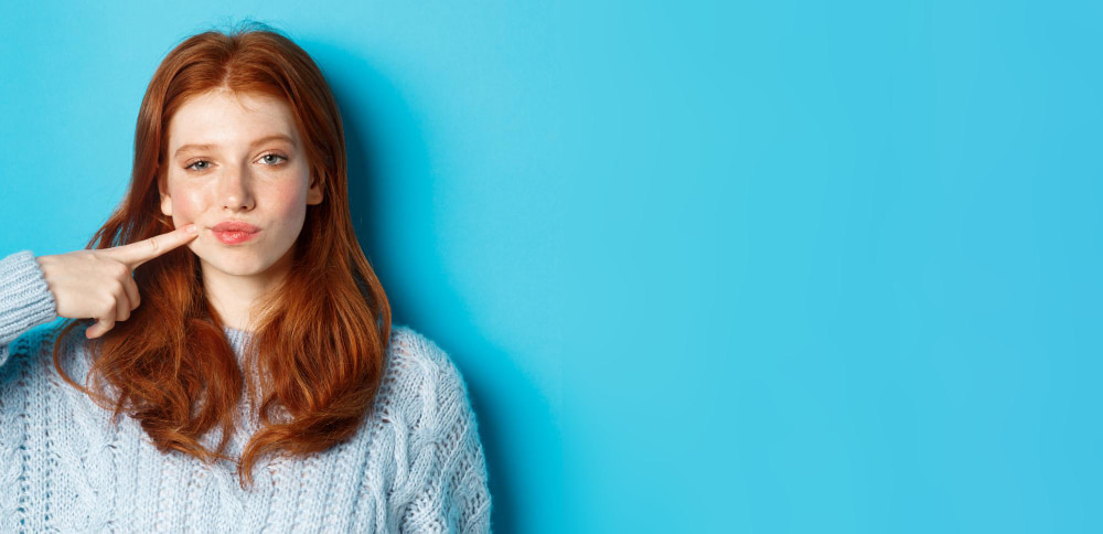 A woman with long red hair in a light blue sweater stands against a bright blue background. She playfully pouts while pointing at her cheek.