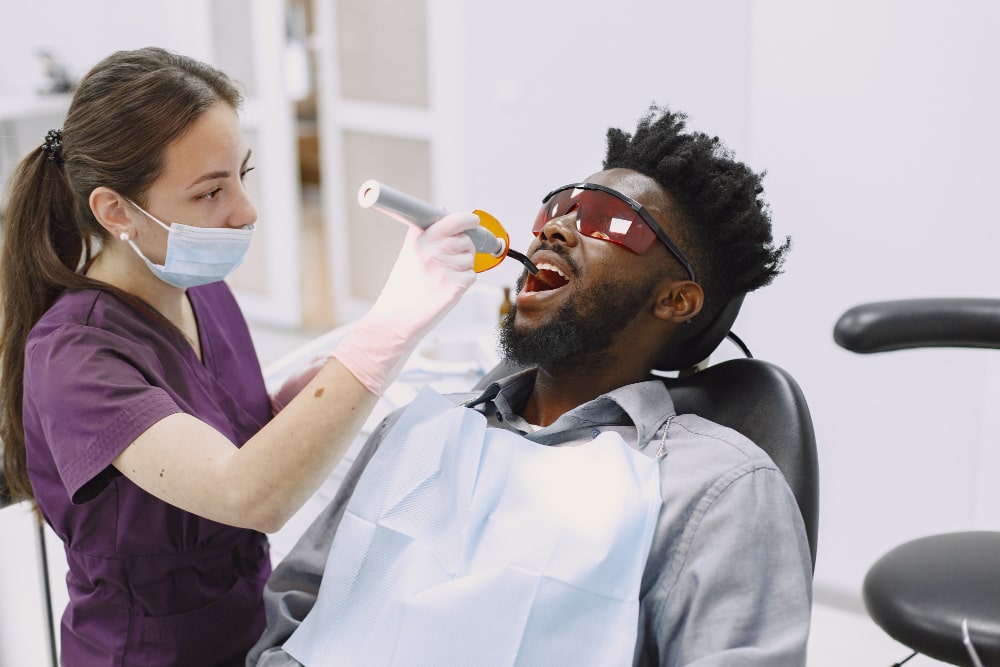 A dentist in purple scrubs and mask uses a dental curing light on a patient wearing protective glasses.