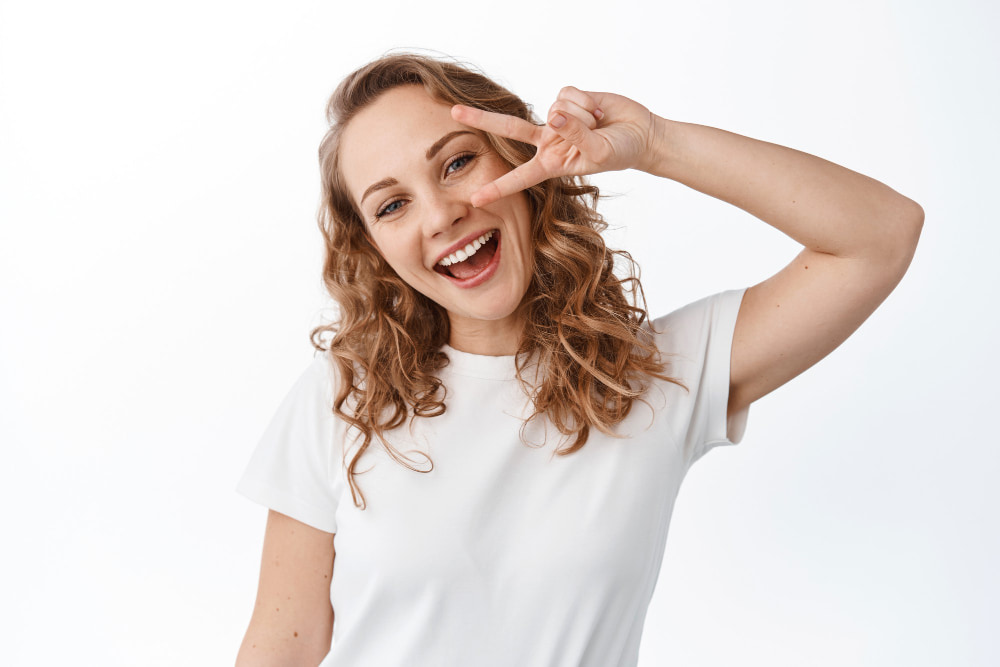 A smiling woman with curly hair shows a playful peace sign near her eye. She wears a white t-shirt against a plain background.