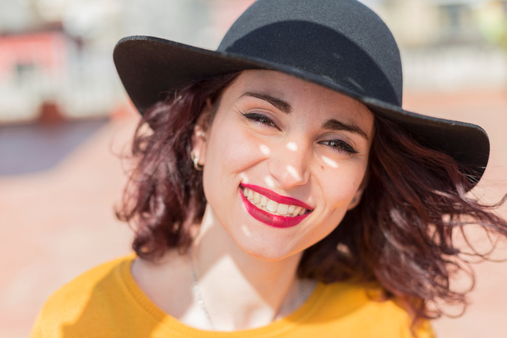 Smiling woman wearing a black hat and yellow top stands outdoors. Her hair is softly blowing in the wind, and sunlight creates playful shadows on her face.