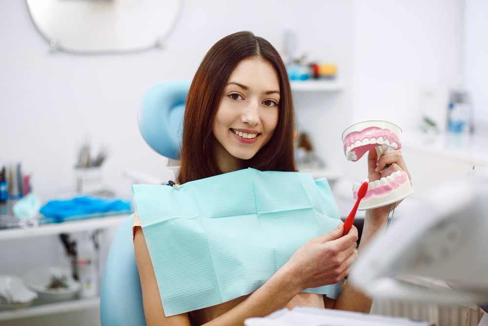 A woman in a dental chair holds a model of teeth and a red toothbrush, smiling. She wears a dental bib, suggesting a clean, educational setting.