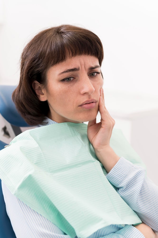 A woman in a dentist chair touches her cheek, appearing concerned and in pain. She wears a dental bib, with a neutral, well-lit background.