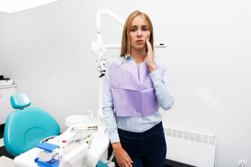 A woman in a dental office looks concerned, holding her cheek. She wears a bib and stands beside dental equipment,