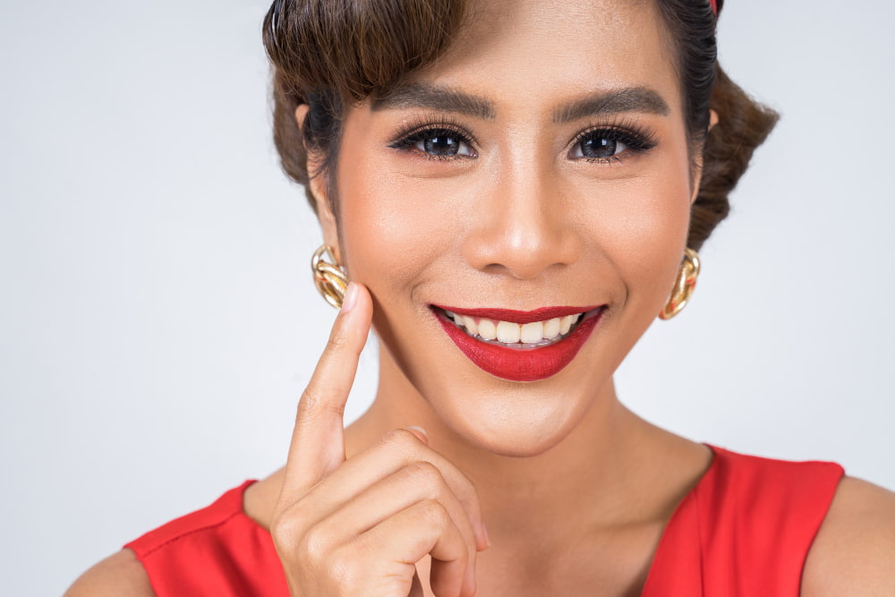 Smiling person wearing red lipstick and gold earrings, with a hand gently touching their cheek.