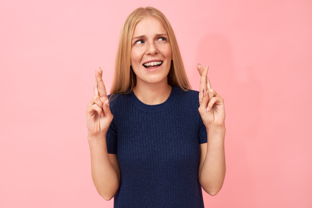 A woman with long blonde hair in a blue shirt stands against a pink background, smiling with fingers crossed.