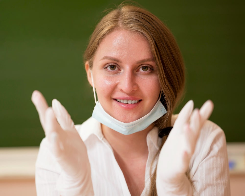 Smiling woman in white blouse, wearing gloves, and a lowered face mask, set against a green background.