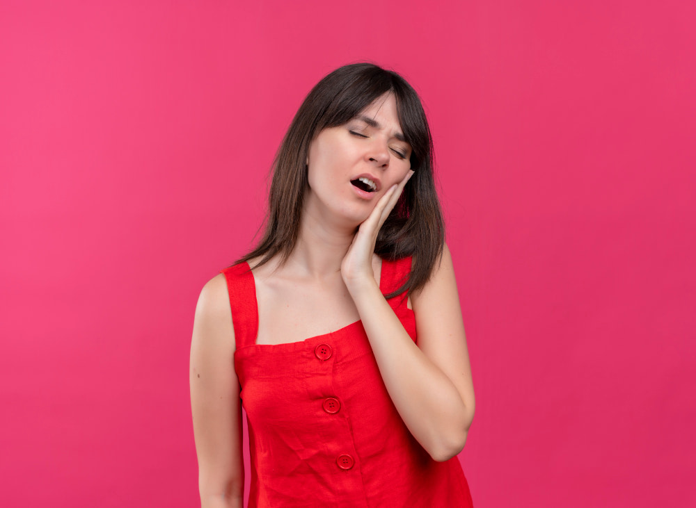 A woman in a red dress stands against a pink background, eyes closed, and holding her cheek with a pained expression, suggesting discomfort or toothache.