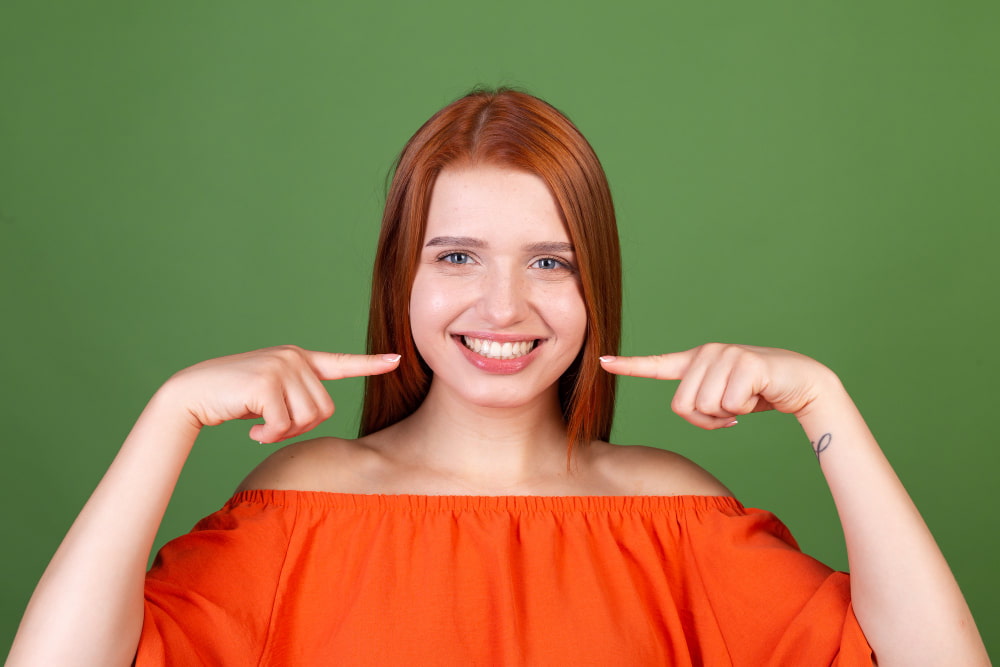 Smiling woman with red hair in an orange top points at her teeth.