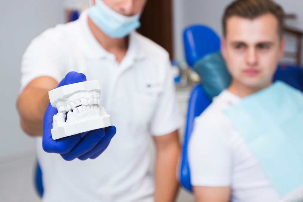 A dentist in gloves holds a dental mold, focusing on it, while a patient in a dental chair watches.