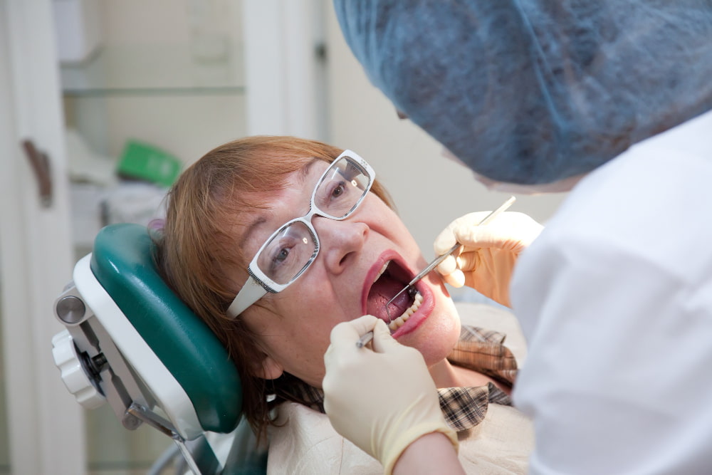 A dentist examines an older patient in a clinic, using dental tools to inspect her mouth.