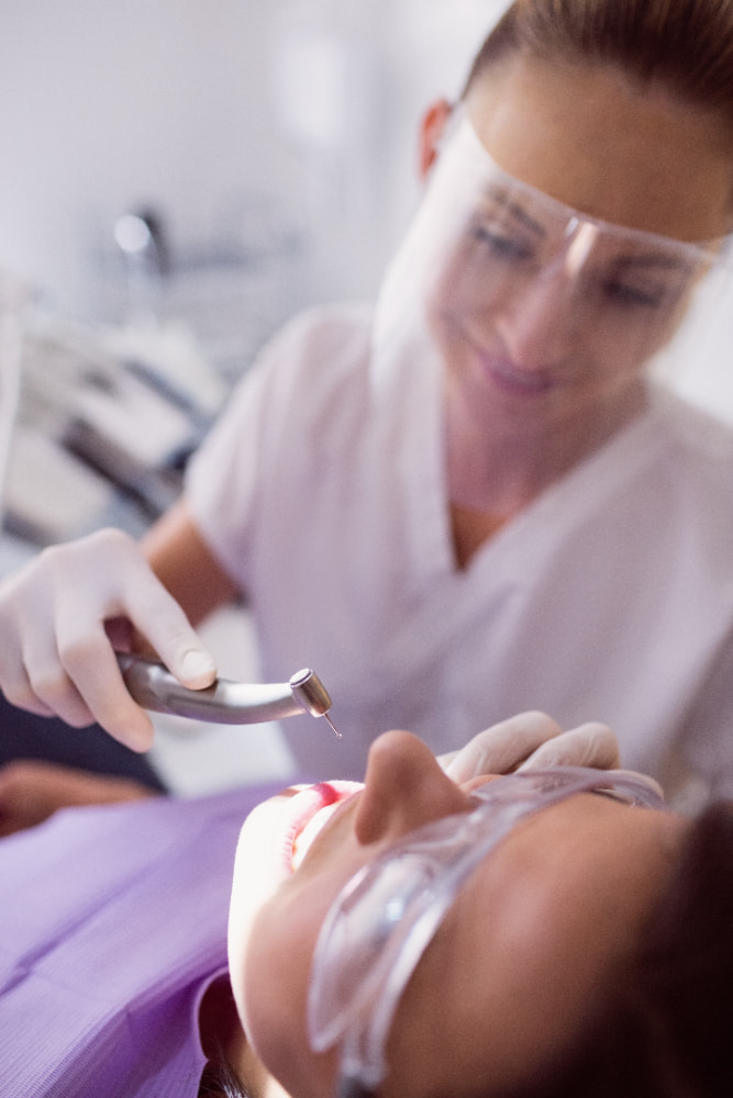 A dentist wearing protective gear works on a patient, who is lying back with safety glasses