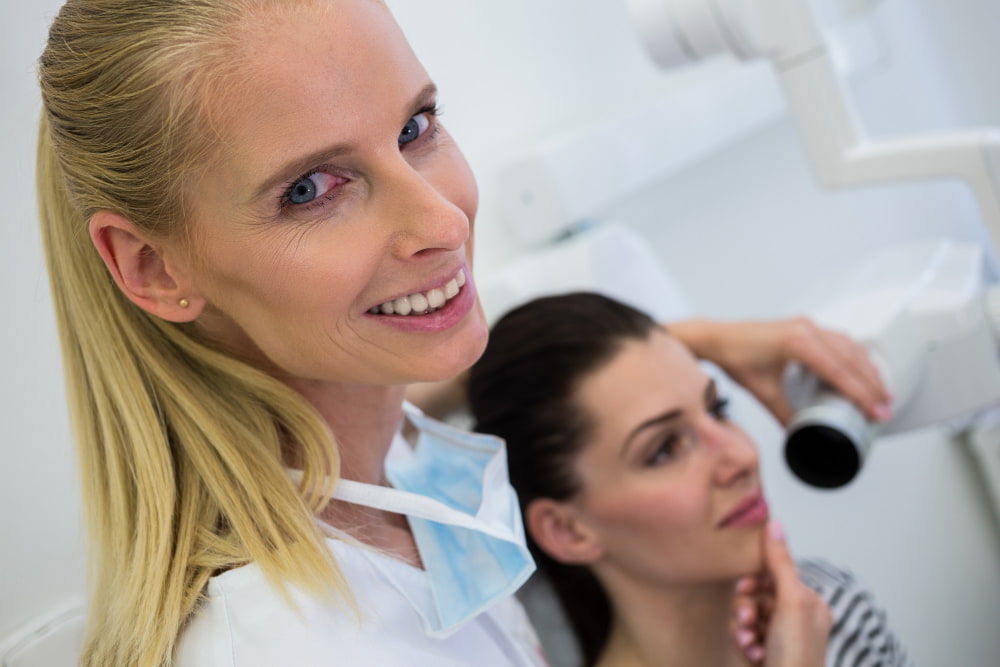 A smiling blonde dentist stands beside a female patient during a cosmetic dentistry.