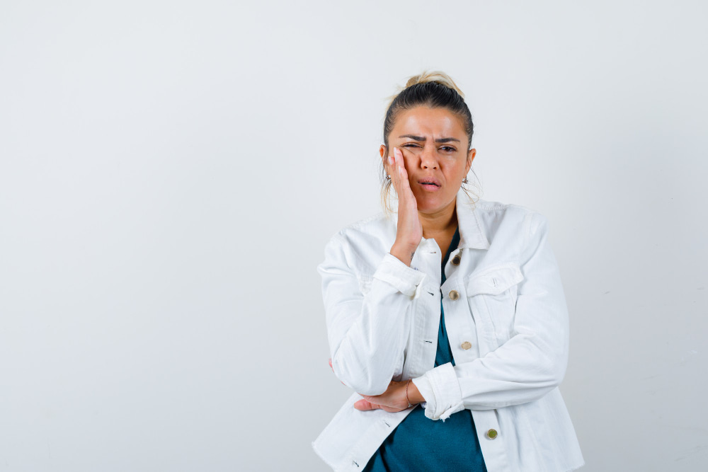 A woman in a white jacket holds her cheek with a pained expression, suggesting discomfort or a toothache,