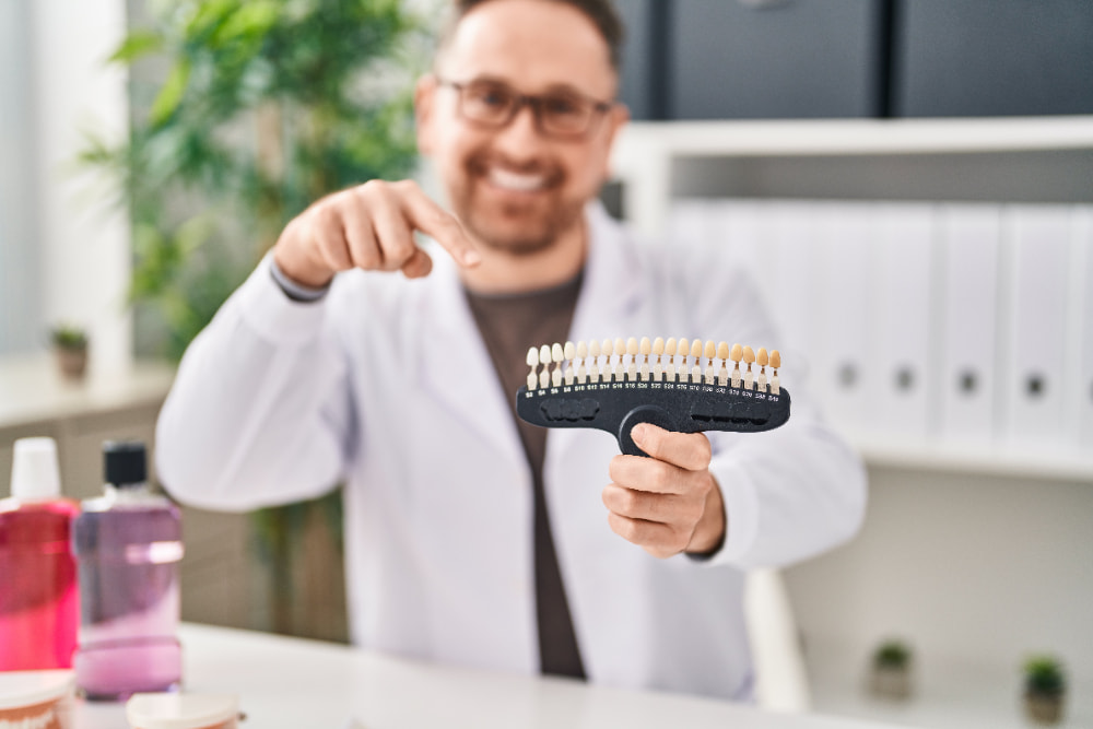 Smiling person in a white coat points to a dental shade guide in an office.