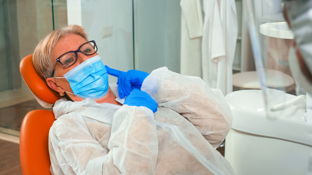 Older woman in a dental chair wearing protective gear, pointing to her cheek.