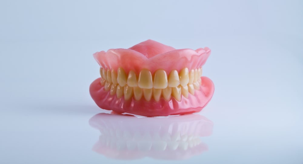 A set of pink dentures with yellow teeth rests on a reflective surface, conveying a sterile tone.