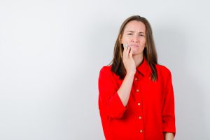 A woman in a red shirt stands against a plain background, touching her cheek with a pained expression.