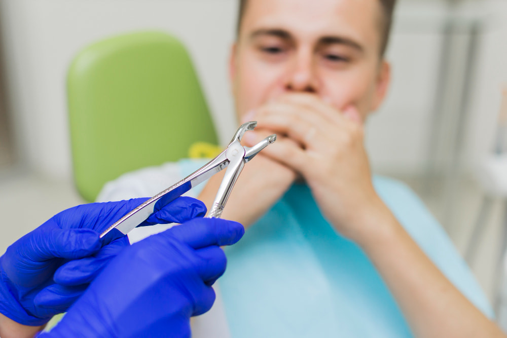 A dentist in blue gloves holds dental forceps, while a patient in a dental chair covers their mouth with both hands,